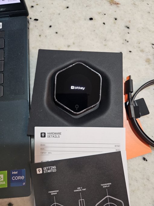 A Bitkey hardware wallet is shown in its packaging alongside a "Getting Started" guide and "Hardware Details" insert, resting next to a laptop and USB-C cable on a granite countertop. The image illustrates the poster's question about calling Bitkey a hardware signing device, since it pairs with a smartphone app rather than generating a traditional seed phrase.