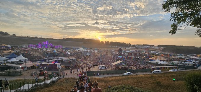 An aerial sunset view of a large outdoor music festival with multiple stages, crowds, and tents, resembling Glastonbury.