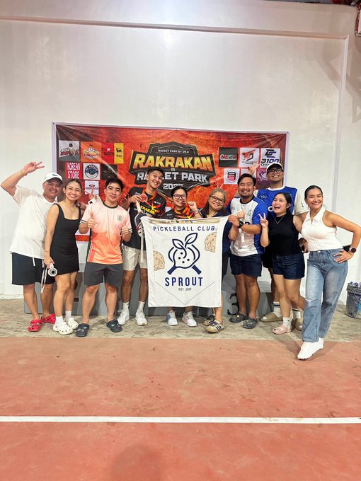A group of people posing on a tennis court holding a banner for 'SPROUT PICKLEBALL CLUB' in front of a 'RAKRAKAN RACEET PARK' event banner, with some wearing medals.