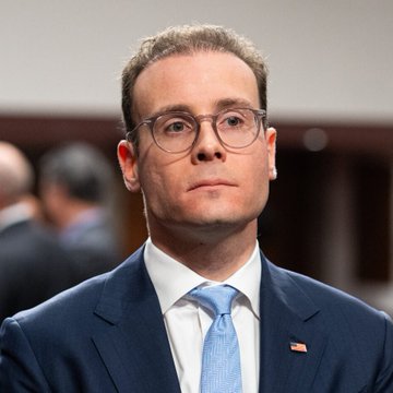 A portrait of a man with short brown hair wearing glasses, a dark blue suit, a white shirt, and a light blue tie, with a small American flag pin on his lapel.