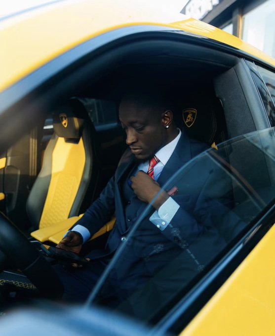 The image shows a man wearing a suit, sitting inside a car with his arm resting on the steering wheel. He appears to be focused on something outside the frame, possibly checking or adjusting his phone. The car has a yellow exterior, which stands out against the background of the image.
