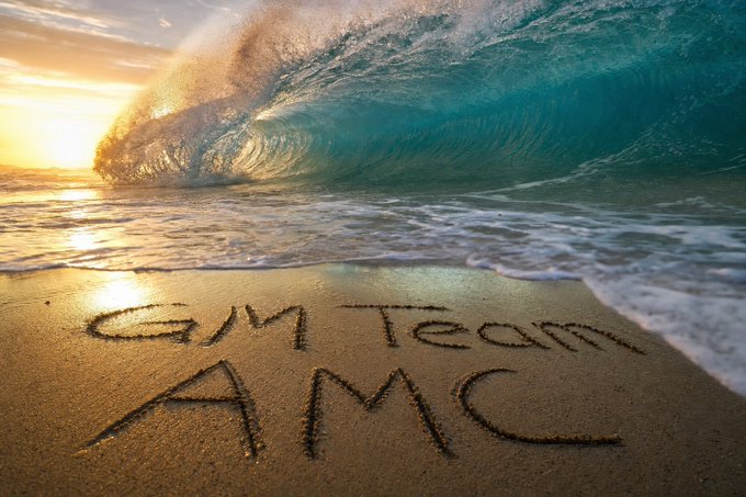 The words 'GM Team AMC' are written in the sand on a beach, with a large turquoise wave crashing under a golden sunset sky.