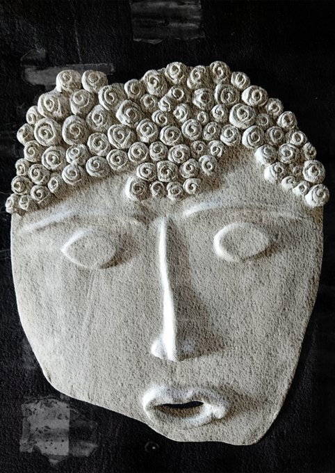 A stone-carved face mask with closed eyes, a long nose, and curly hair decorations is mounted on a dark surface.