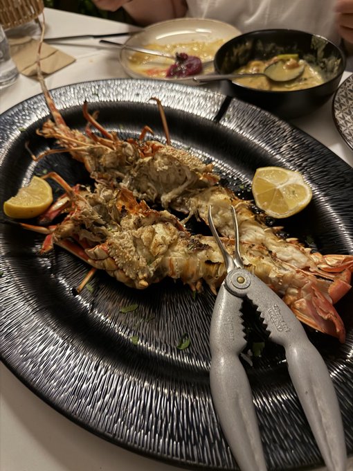 A cooked lobster is presented on a large textured black plate with lemon wedges, accompanied by seafood crackers and side dishes of butter and sauce on the table.