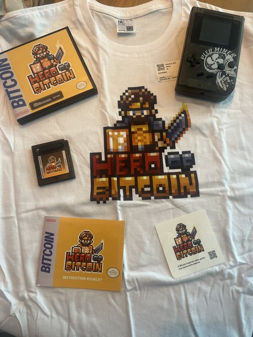 A collection of 'HERO BITCOIN' merchandise is displayed on a white t-shirt, including the shirt itself featuring a pixel-art knight graphic, a Game Boy-style handheld console, a game cartridge, an instruction booklet, and a card, all bearing the 'BITCOIN' branding.