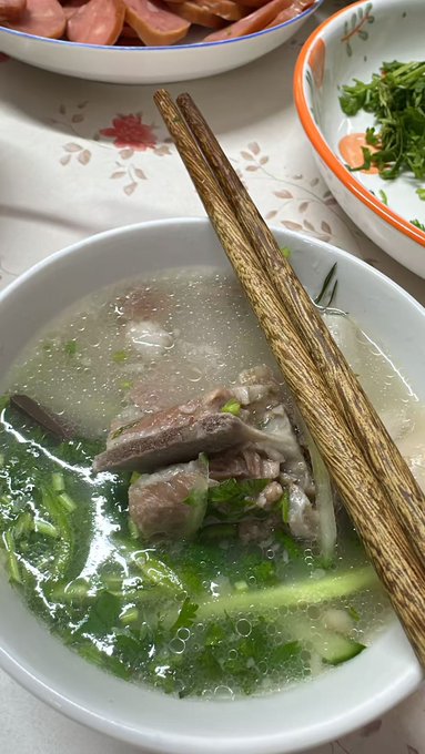 A bowl of clear Vietnamese soup, likely phở, containing meat and green herbs, with wooden chopsticks resting across the top.