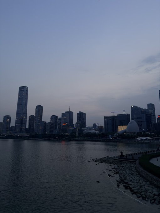 Twilight cityscape over a river, showing a skyline of skyscrapers, a domed building, and a person standing on a rocky shoreline.