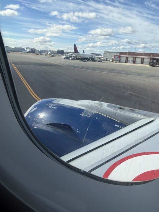 A view from an airplane window showing the blue engine and tarmac, with another aircraft (featuring a Delta tail) visible at a gate in the background under a partly cloudy sky.
