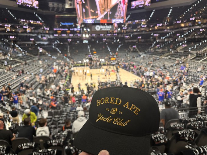 The image shows a person wearing a black baseball cap with gold lettering that reads "Bored Ape Yacht Club". The hat appears to be tilted, as if someone has just put it on or taken it off. In the background, there is a large crowd of people gathered in what seems to be an arena or stadium setting.

The caption above the image provides additional information about the hat and its connection to the "Bored Ape Yacht Club" event.