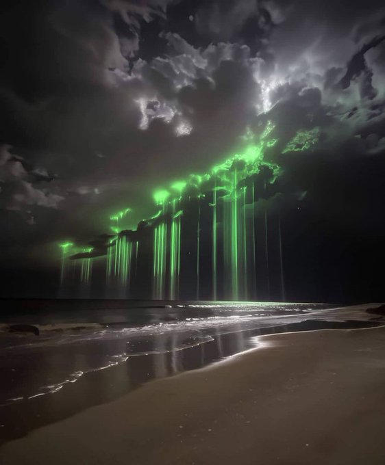 A nighttime beach scene with spectacular green luminous light streaks appearing to fall from dark stormy clouds onto the shoreline, creating haunting reflections on the wet sand.