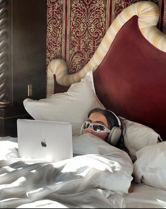 The image shows a woman lying down on her bed with an open laptop computer next to her. She appears to be wearing headphones and sunglasses while using the laptop. The background of the image features a red headboard, which adds a pop of color to the scene. The caption provided in the image seems unrelated to the main subject matter and does not provide any additional context or information about the content displayed on the laptop screen.