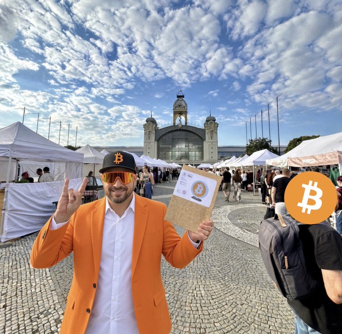 A man in an orange blazer, white shirt, Bitcoin-themed cap, and reflective sunglasses holds up a Bitcoin certificate at an outdoor crypto event, with a Bitcoin logo sticker visible.
