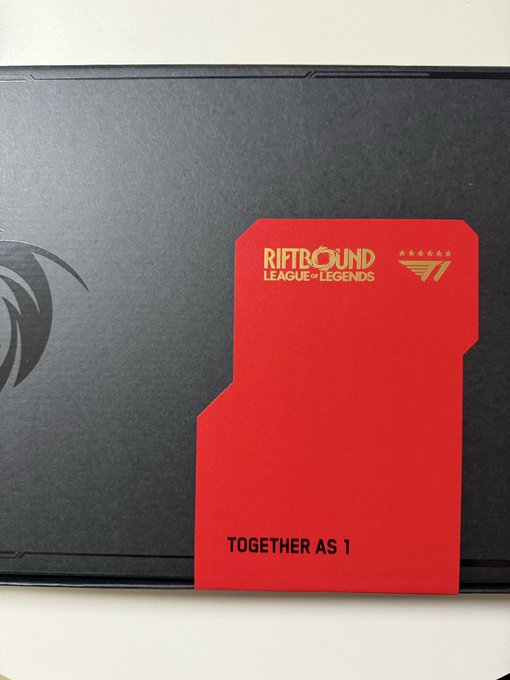 A red T1 Riftbound League of Legends themed card reading 'TOGETHER AS 1' rests against a black laptop featuring a T1 logo.