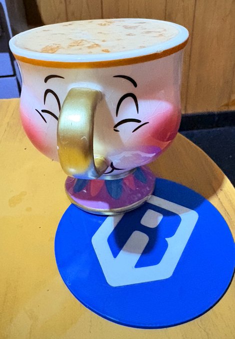 A whimsical ceramic mug shaped like a smiling face with rosy cheeks, gold handle, and decorative purple base, sitting on a blue coaster with a white logo.