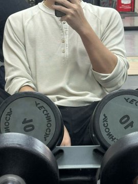 A person in a cream-colored henley shirt sits on a gym bench holding a phone, with two 10 lb Technogym dumbbells visible in the foreground.