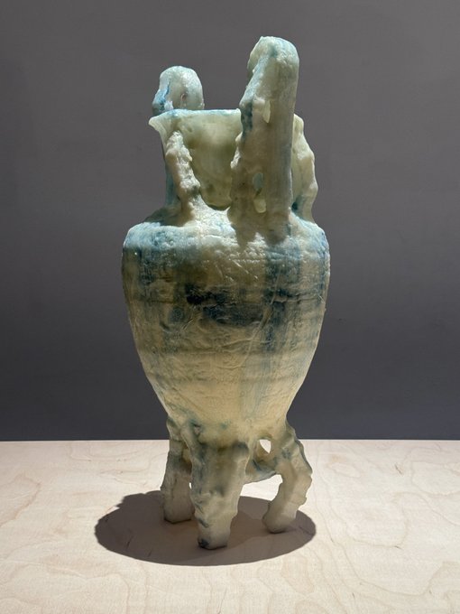 A sculptural vessel with rough, organic textures in cream and greenish-blue glazes, featuring two tall handles and a tripod base, displayed on a light wooden surface against a gray background.