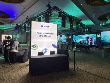 A dimly lit event space features a Trust Wallet booth with a brightly lit sign reading "TRUST" and "The crypto wallet you control," illuminated against a backdrop of colorful stage lighting, matching the post's description of Trust Wallet partnering with Wanwan and Hyperliquid at the event.
