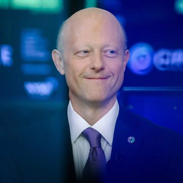 A close-up portrait of a smiling bald man in a dark blue suit and purple tie, photographed at what appears to be a formal corporate or technology event with blue-lit screens in the background.