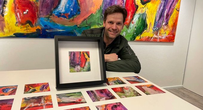 A smiling man in a green jacket sits at a table displaying colorful abstract artworks, holding a framed abstract piece, with a larger abstract painting hanging on the wall behind him.