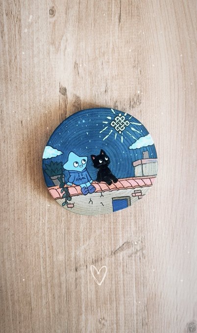 A round hand-painted enamel pin showing a blue cat and black cat on a rooftop under a starry night sky, on a wooden surface.