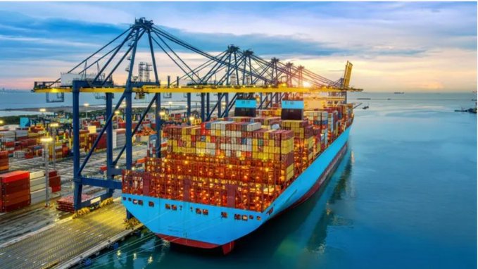 An aerial view captures a massive blue MSC container ship docked at a port at dusk, loaded with colorful shipping containers and illuminated by bright orange crane lights.