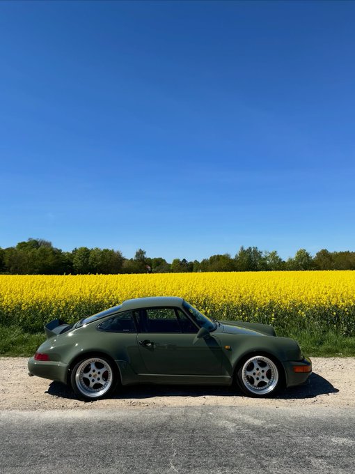 A dark green Porsche 911 parked on a road beside a vibrant yellow field under a clear blue sky.