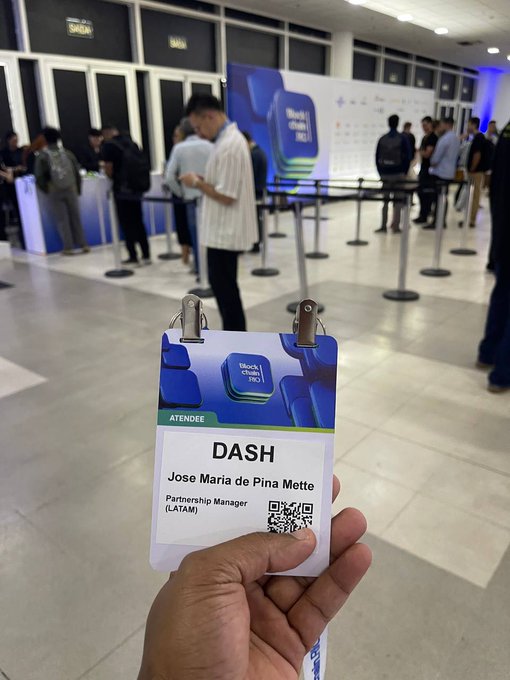 A hand holds up a "DASH" attendee badge for Jose Maria de Pina Matte, Partnership Manager (LATAM) at Blockchain Rio, with registration desks and networking crowds visible in the background of the conference venue. The badge ties directly to the post's mention of productive Day 1 conversations focused on new Dash integrations and expanding local crypto utility in Latin America.