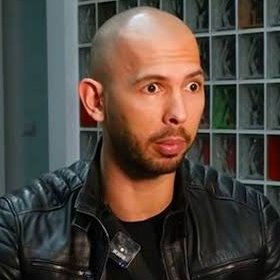 The image features a bald man wearing a leather jacket with an electronic device attached to his neck. The device appears to be a microphone or a camera, suggesting that he might be involved in some form of media production or broadcasting activity. The background shows a bookshelf filled with various books and DVDs, indicating that the man could be working from home or in a studio setting where he needs access to reference materials for his work.
