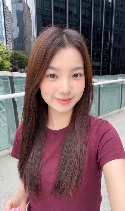 A young woman with long brown hair wearing a maroon t-shirt takes a selfie outdoors, with modern glass skyscrapers and a railing visible in the background.