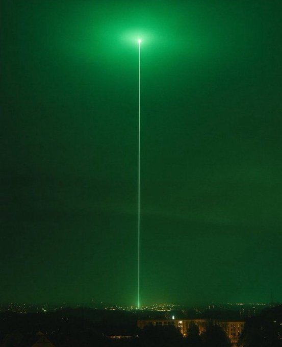 A green laser beam shoots straight up from a city skyline into the night sky, topped with a bright light, against a dark green atmospheric background.