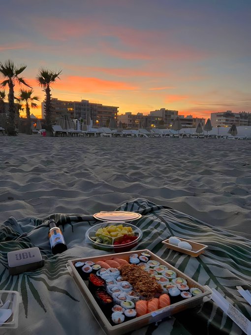 A beach scene at sunset featuring a striped blanket with a large sushi platter, fruit, drinks, and a JBL speaker, with lounge chairs and palm trees in the background.