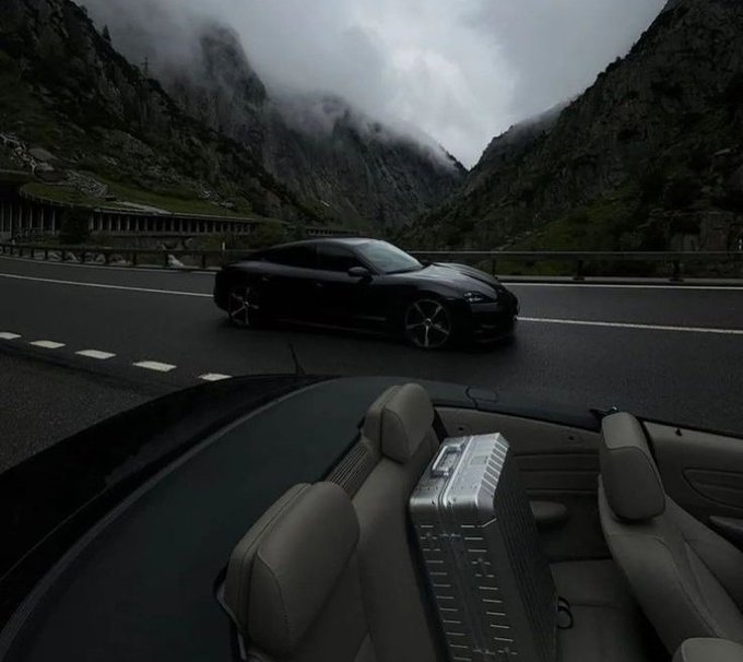 A black sports car drives on a wet mountain road through a foggy valley, with the interior of another convertible car visible in the foreground.