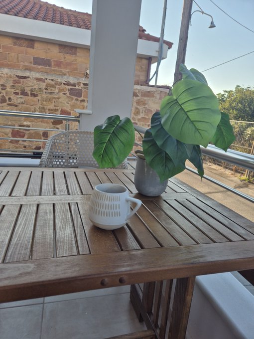 A serene balcony scene with a wooden table holding a white ceramic mug and a potted monstera plant, set against a backdrop of a brick wall and railing.