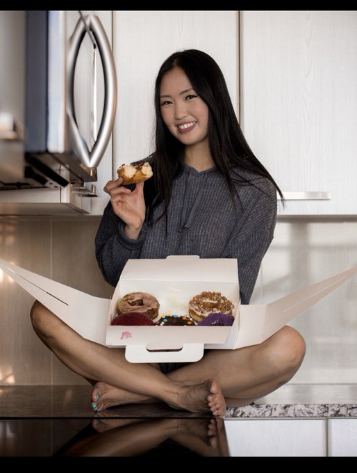 A smiling young woman sitting cross-legged on a kitchen counter, holding a partially eaten pastry with an open box of assorted donuts in front of her.