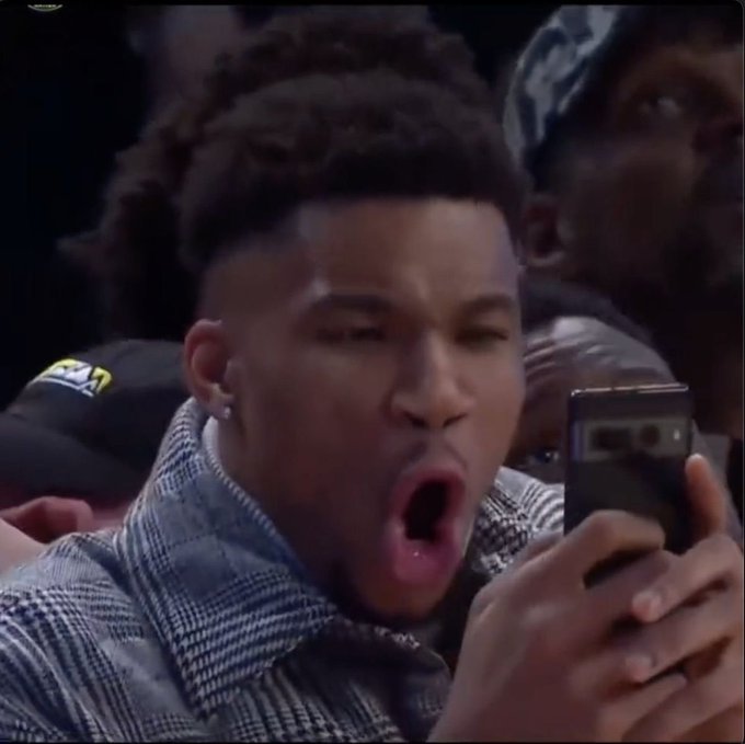 A young man with a dreadlock fade and an earring holds up a smartphone, his mouth wide open in apparent shock, while wearing a checkered gray shirt at what appears to be a sporting event. The image humorously captures the "red candles" meme reaction to the failed CLARITY Act vote, depicting the stunned disbelief investors feel watching crypto prices plummet.