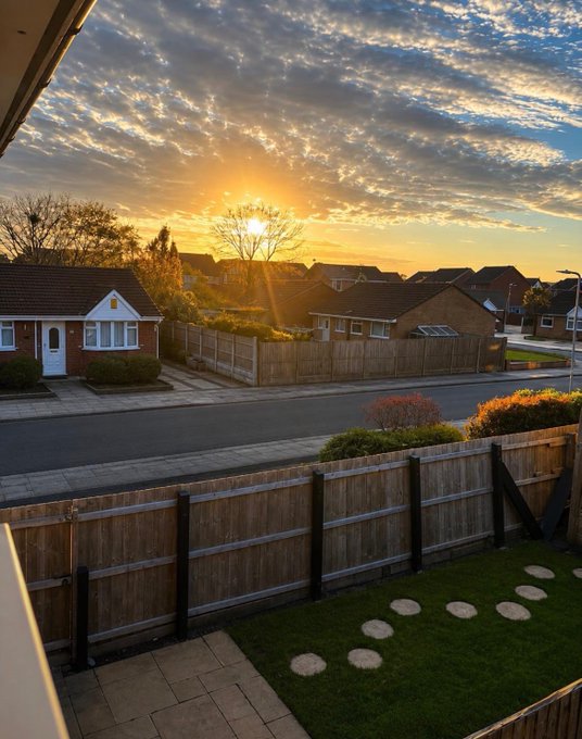 A serene sunrise over a residential neighborhood, viewed from a backyard with wooden fencing and stepping stones on the lawn.