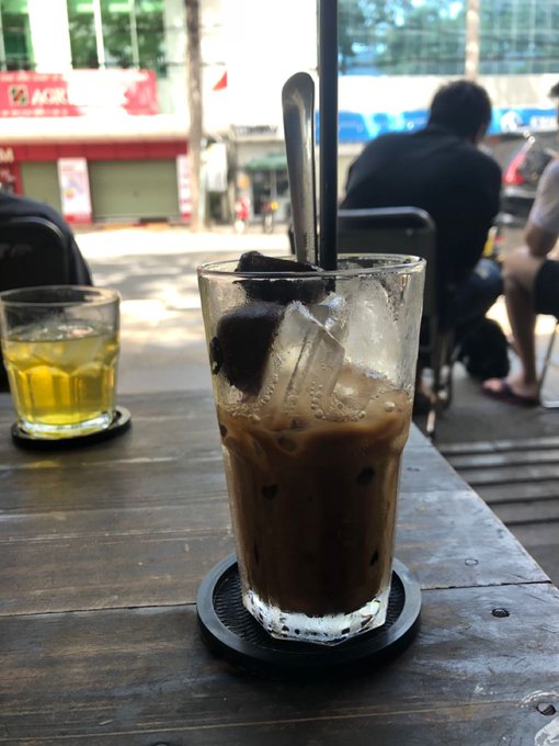 A glass of iced coffee with a spoon resting in it sits on a dark wooden table next to a smaller glass of a yellow beverage, taken at a street-side cafe with people and a storefront in the background.