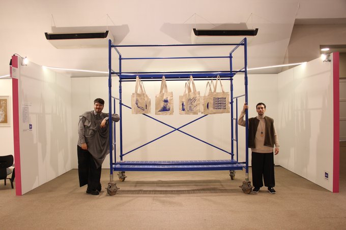 Two men stand beside a blue metal scaffolding displaying hanging tote bags at the "What's Happening on the Internet?" exhibition, curated by @stipinpixel for @maskaygallery in partnership with Art on Tezos at the Tucumán Contemporary Art Fair.