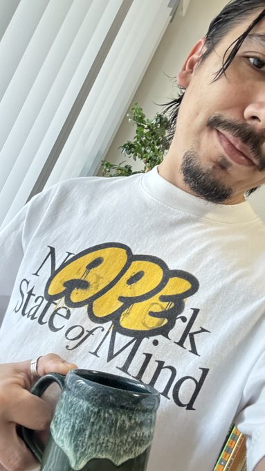 A person with long dark hair and a goatee wearing a white 'N*** State of Mind' t-shirt with 'GG' logo, holding a dark ceramic mug in front of vertical blinds and a plant.