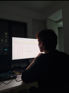 Person viewed from behind, sitting in a dimly lit room looking at a computer monitor displaying a spreadsheet with colorful rows.