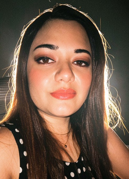 A selfie-style portrait of a young woman with long dark hair, wearing a black and white polka dot top and a delicate necklace, photographed against a dim background with warm lighting.