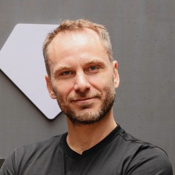A portrait of a man with short hair and a beard wearing a black shirt, standing in front of a gray wall.