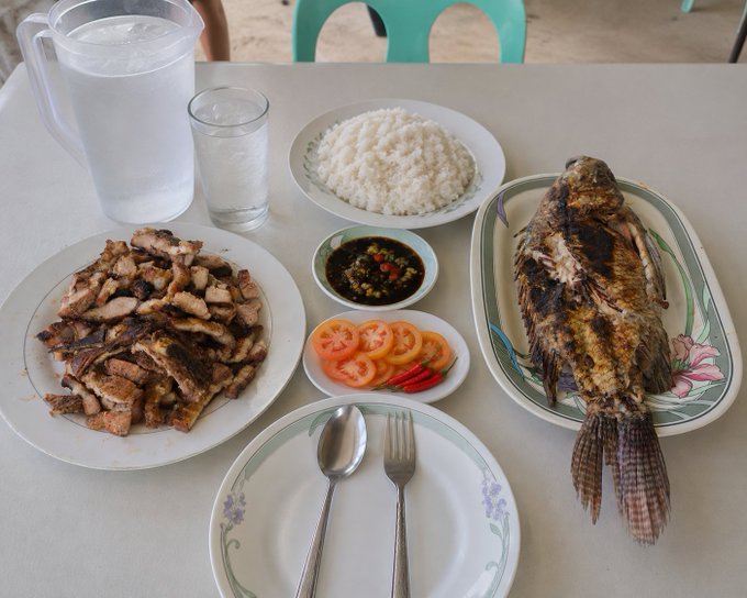 A spread of food on a table featuring grilled pork, a whole grilled fish, white rice, sliced tomatoes, chili peppers, and a dipping sauce, with a pitcher of water.