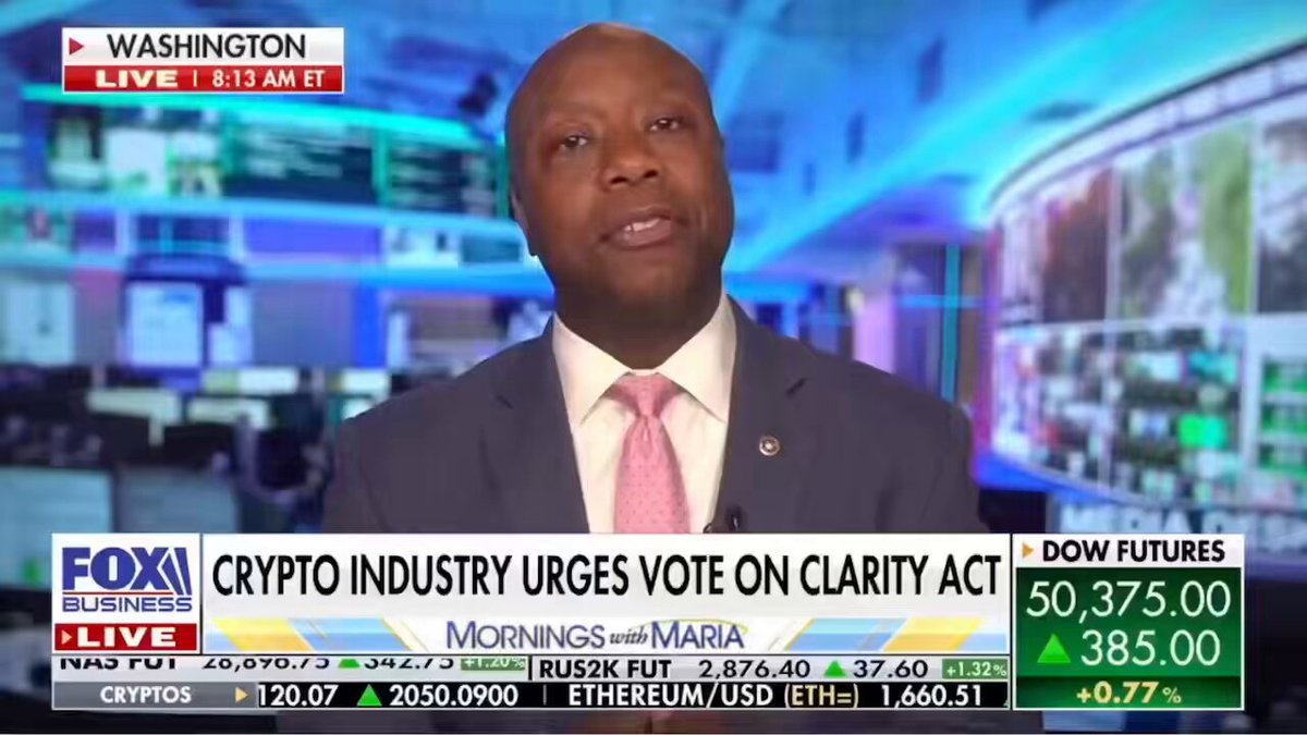 The image shows a man wearing a suit, sitting at a desk with a computer screen behind him. The news anchor is reporting about cryptocurrency and its impact on the financial industry. The man appears to be discussing the failures of the clarity act and how banks are adopting Bitcoin as their future currency.
