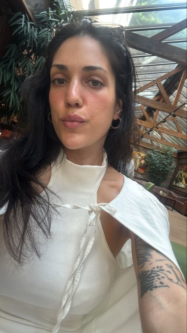 A dark-haired woman with a nose ring and a tattooed arm takes a selfie wearing a white cut-out top.