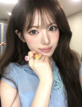 A young woman with long brown hair poses for a selfie, wearing a light blue top and colorful gemstone rings.
