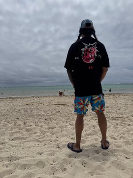 A man stands on a sandy beach under an overcast sky, wearing a black t-shirt with a red graphic, blue patterned shorts, and a cap, facing the ocean.