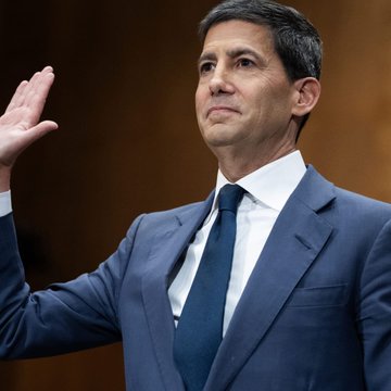 A man in a blue suit and tie raises his right hand as if taking an oath, set against a dark wood-paneled background.