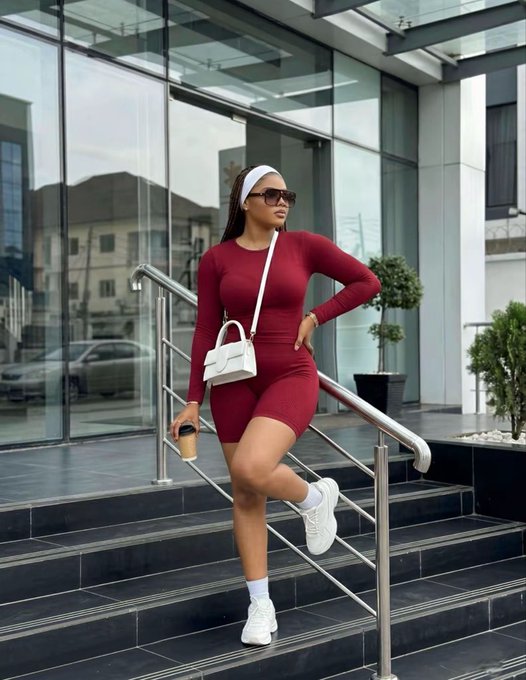 A woman in a maroon long-sleeve bodycon dress and sunglasses posing on outdoor steps while holding a coffee cup and a white handbag.