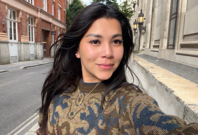 A selfie of a young woman with long dark wavy hair wearing a camouflage-pattern sweater, taken on a street with classic brick buildings in the background.